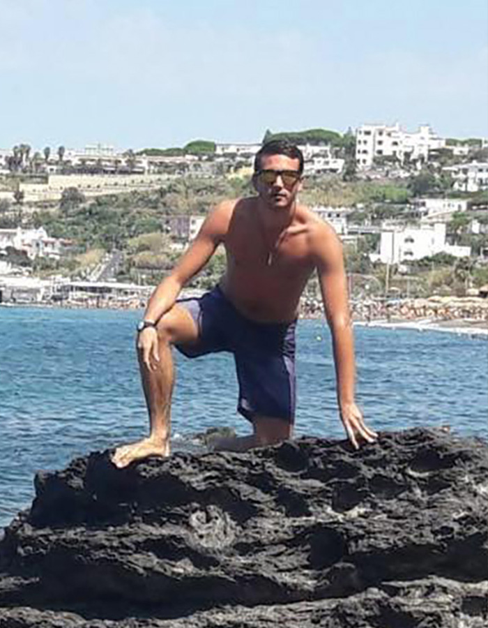 Homem posando sem camisa na costa rochosa com mar e edifícios ao fundo