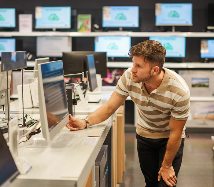 A man in a striped polo shirt looking at a computer monitor in an electronics store. This relates to expensive habits that save money.