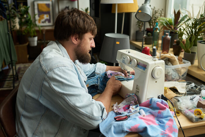 Man with a beard sewing jeans, demonstrating a habit to save you money.