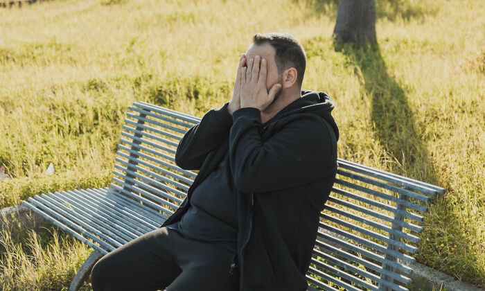 A man sits on a park bench with his face in his hands, looking distraught, conveying the need for Routine Cancer Screening.