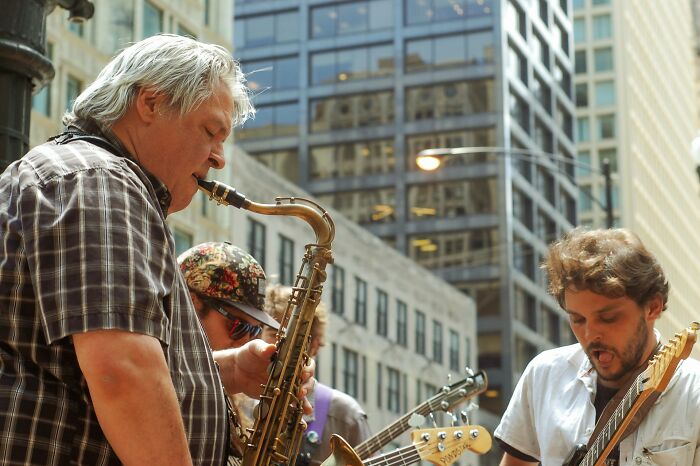 Two musicians performing on a city street, one playing saxophone, the other an electric guitar. Celebrity relatives.