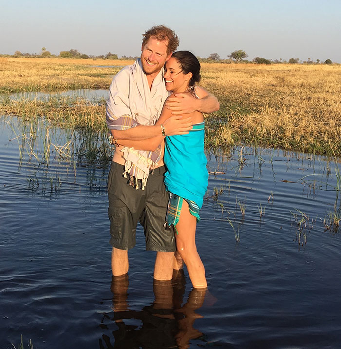 Meghan Markle and Prince Harry embrace in water. Meghan is wrapped in a blue sarong, Harry in a white shirt, both smiling.