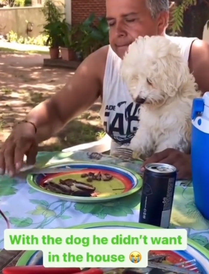 A man, initially against getting a pet, now lovingly holds a small white dog at a table, demonstrating Men Got A Pet Against Their Will.