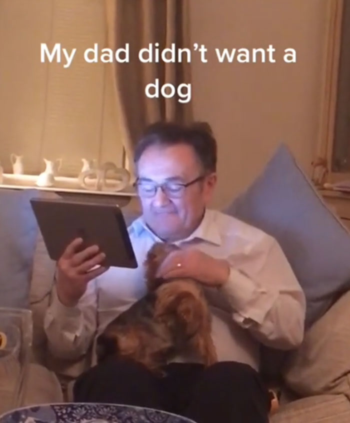 A man, initially reluctant to get a pet, happily pets a small dog on his lap while looking at a tablet.