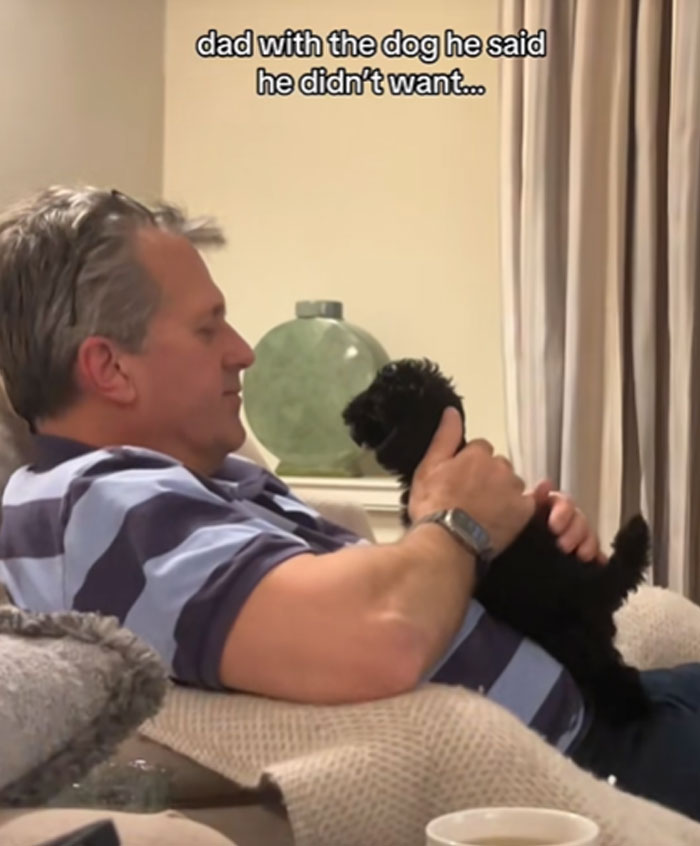 A man in a striped shirt gently holding a black puppy, illustrating men getting a pet against their will.