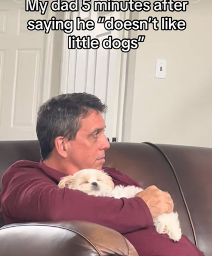 A man in a red shirt cuddling a small white dog, a perfect example of men getting pets against their will.