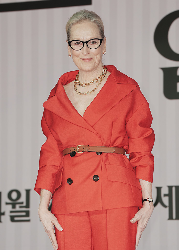 Meryl Streep, wearing glasses and a red suit, smiling. A celebrity potentially skipping the Met Gala.