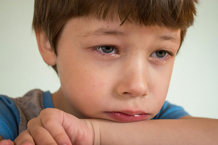 A young boy with brown hair and tearful eyes rests his head on his arm. Messed up school stories can be hard.