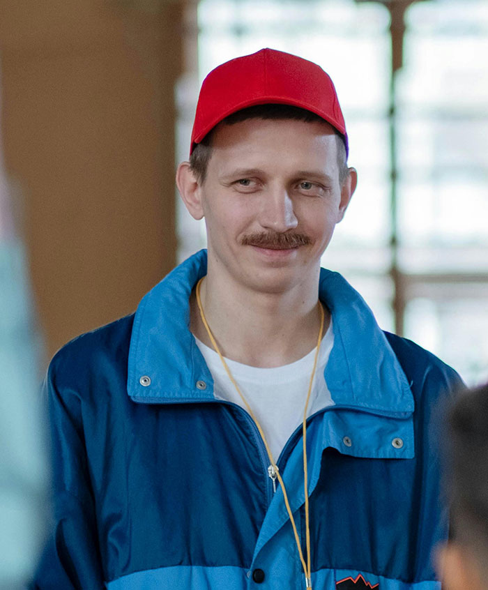 A smiling man in a red cap, blue jacket, and yellow lanyard, hinting at messed up school stories.