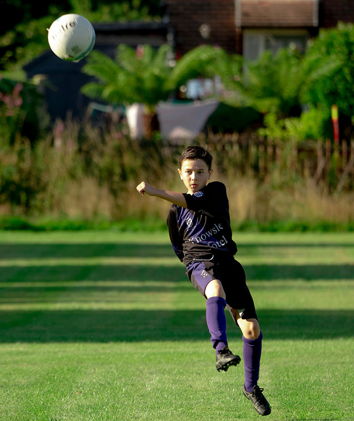 A young boy playing soccer, mid-air, kicking a ball. Reminiscent of younger years and school stories.