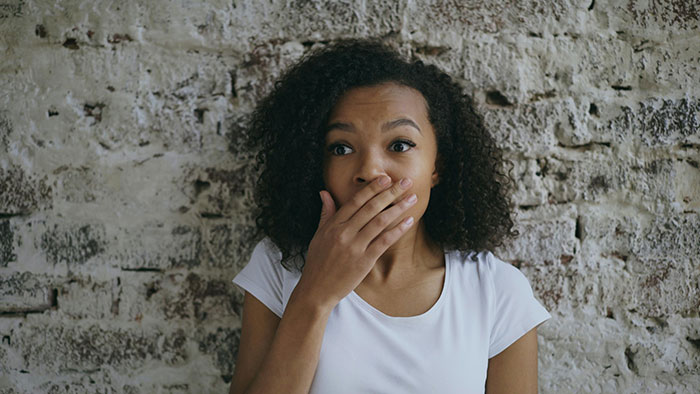 A young woman with curly hair covers her mouth in shock against a brick wall, reflecting messed up school stories.