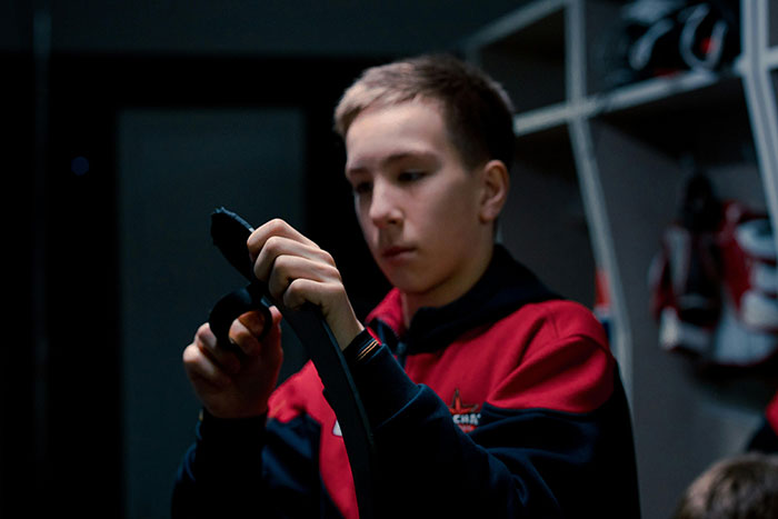 A young boy in a locker room, focusing on wrapping a hockey stick, reflecting school stories.