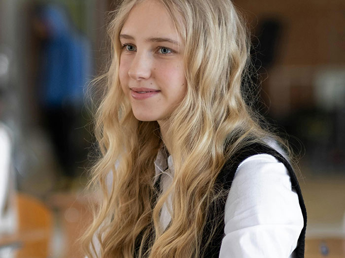 A young blonde student with wavy hair smiling gently, reflecting on school stories and younger years.