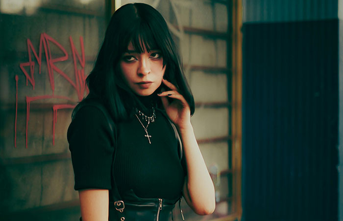 A young woman with black hair and bangs, wearing a black top and cross necklace. Messed up school stories.