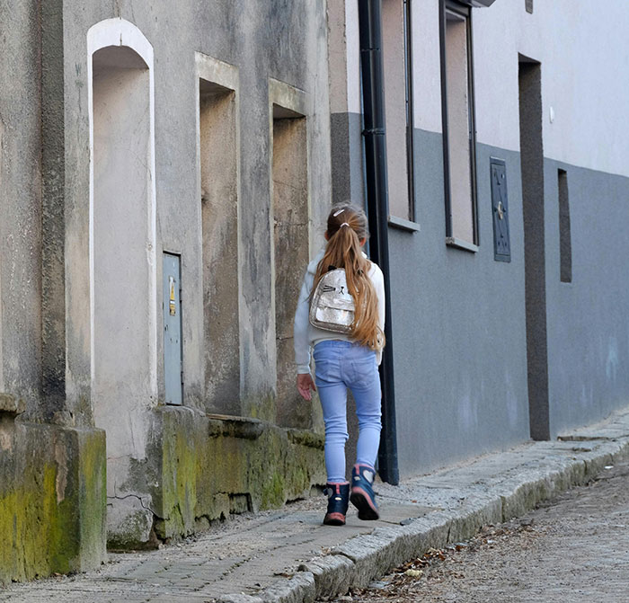 A young girl with a backpack and long hair walks down a street, evoking school stories and younger years memories.