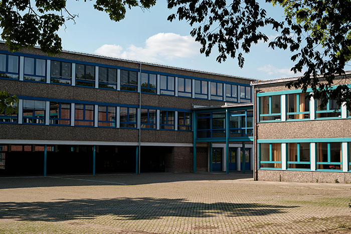 A concrete school building with numerous blue-framed windows and a large paved courtyard, hinting at messed up school stories.