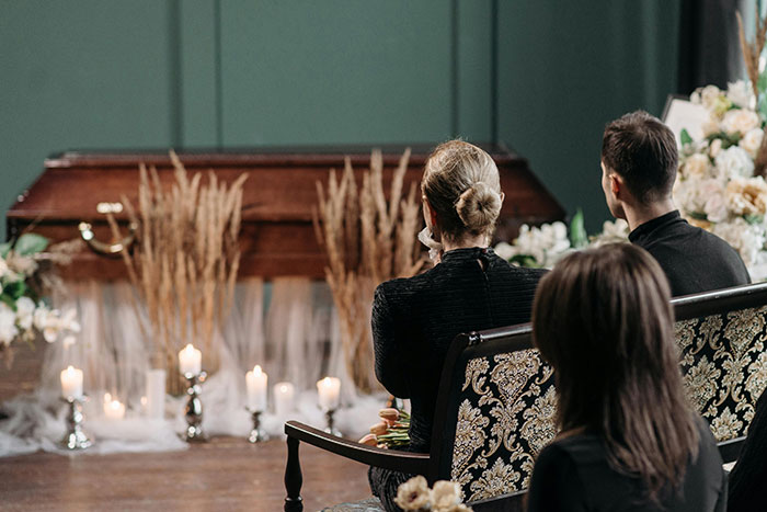 Mourners at a funeral service, a coffin decorated with dried pampas grass. This evokes Messed Up School Stories.