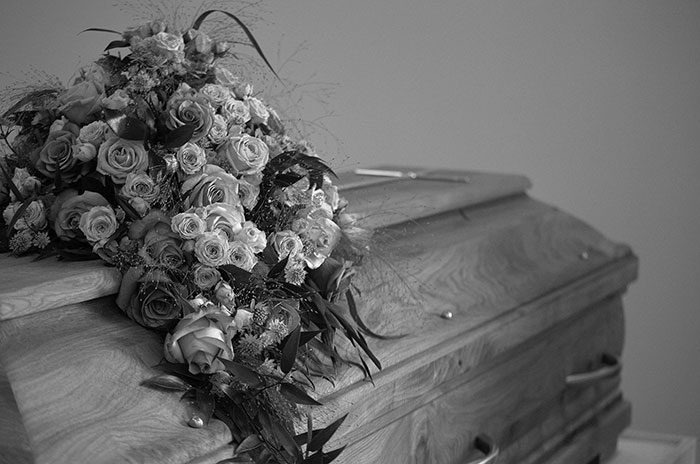 Black and white image of a casket adorned with a large bouquet of roses. A solemn reminder of messed up school stories.