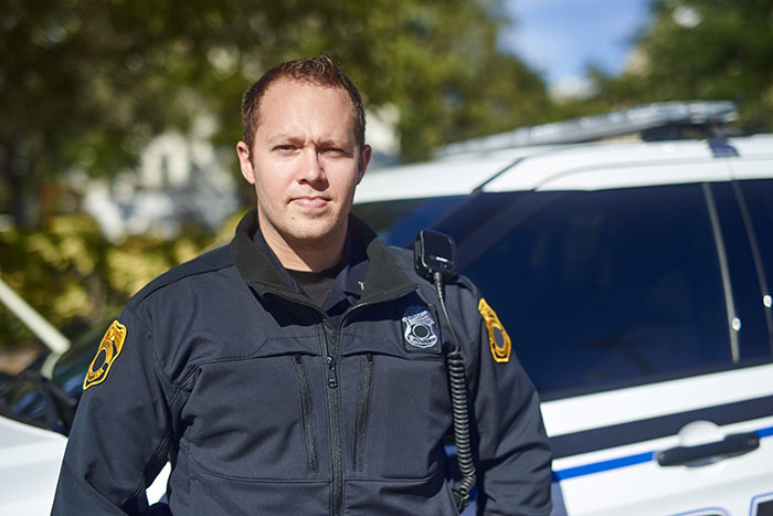 A male police officer in uniform stands in front of a patrol car. He is looking at the camera. mil broke in son.