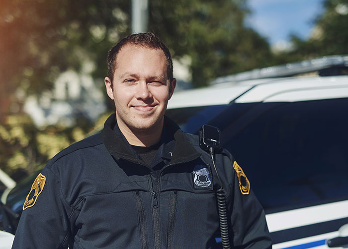 Smiling police officer in uniform standing in front of a patrol car. The officer clarifies how adulthood works.