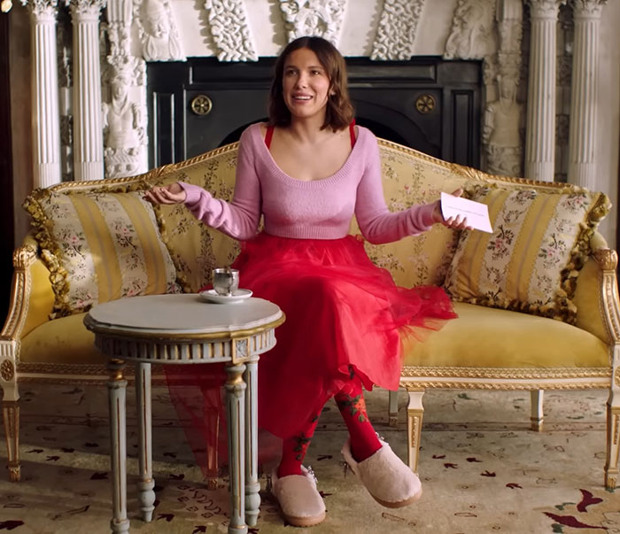 Millie Bobby Brown in pink sweater and red skirt sitting on ornate sofa discussing travel habits Millie Bobby Brown in pink sweater and red skirt sitting on ornate sofa discussing travel habits