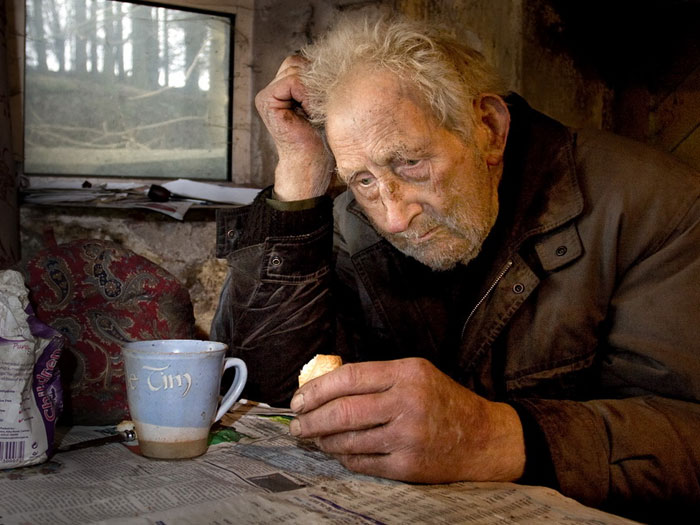Elderly man in distress in a dim room holding bread and sitting at a table with a cup