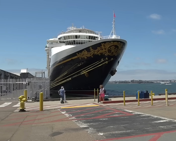 A Disney Cruise ship docked at a port, with people standing on the pier. A large ship and blue sky are visible. Watch your kids.