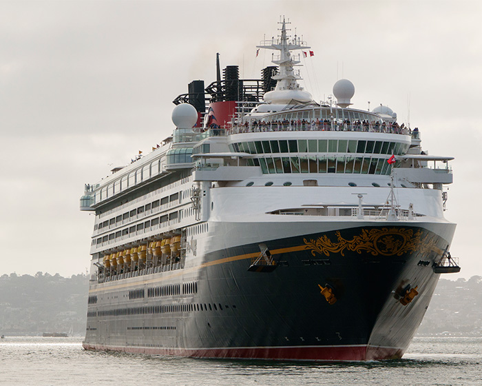 A large Disney cruise ship, featuring Mickey Mouse's colors and gold swirls on its bow, sails on calm water. Watch your kids.