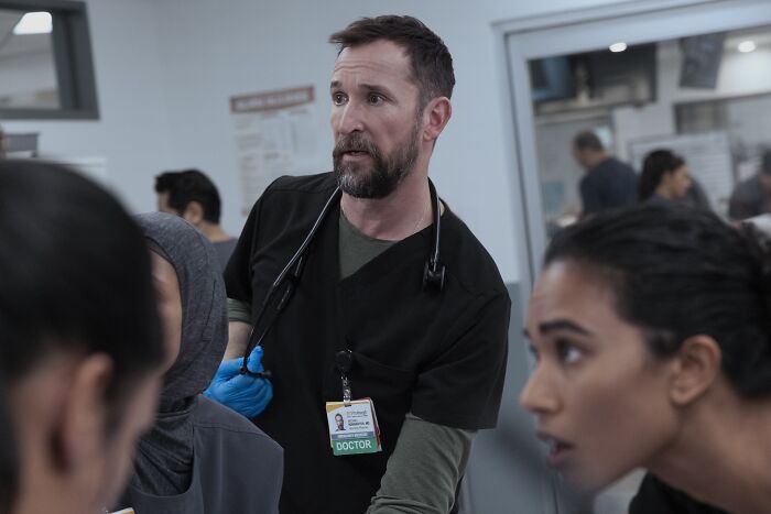 Noah Wyle como médico vestindo uniforme preto e luvas azuis na cena do hospital em The Pitt