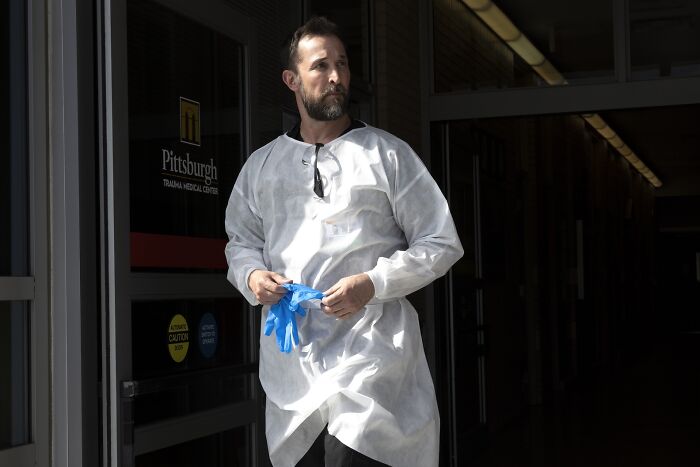 Noah Wyle com bata protetora de hospital e calçando luvas azuis fora do centro médico
