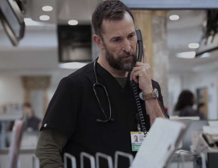 Noah Wyle de uniforme falando ao telefone com estetoscópio em ambiente hospitalar