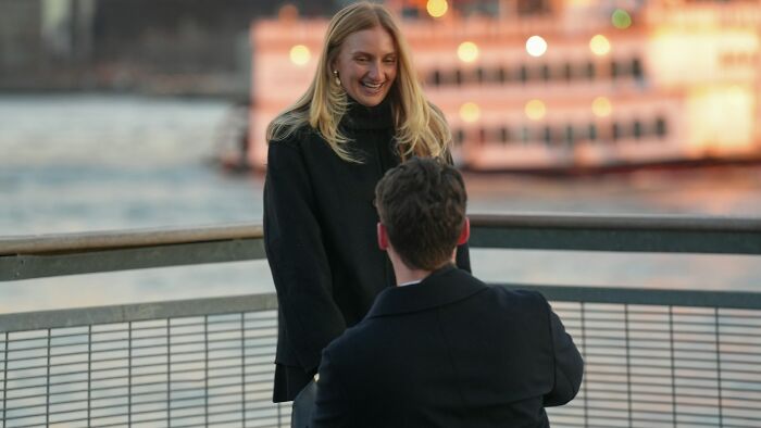 A man proposes to a surprised woman on a pier, with a ferry and city lights in the background. Hilarious engagement stories often begin this way.
