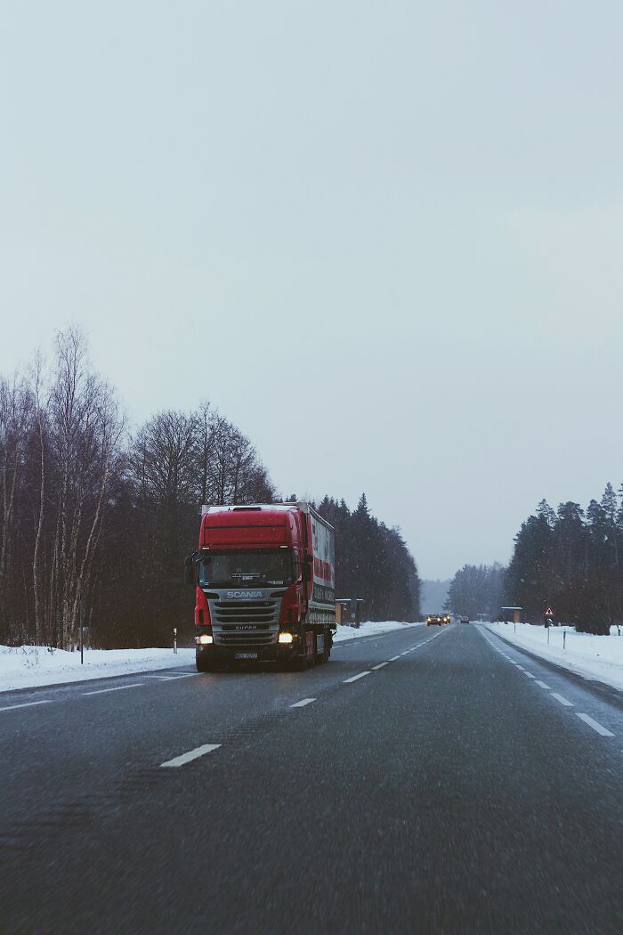 Red truck driving on snowy highway with bare trees on roadside