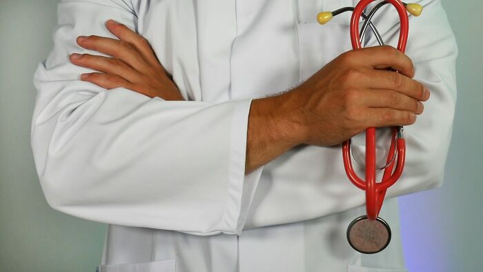 A doctor in a white coat with arms crossed, holding a red stethoscope, symbolizing health and "the other side" of medical stories.