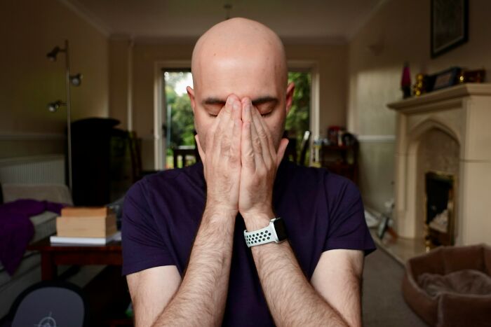 Bald man in a purple shirt covering his face with hands, experiencing creepy things in his living room.