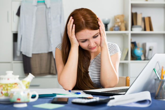 A woman with long brown hair, hands on her head, looking distressed at a table with a laptop, calculator, and papers, feels betrayed about inheritance.