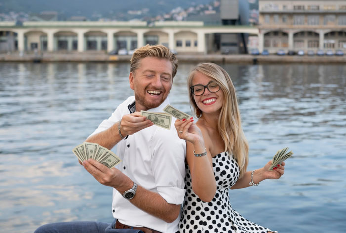 A happy couple holds US dollar bills, smiling near a body of water. This illustrates spending inheritance on France, not a house deposit.