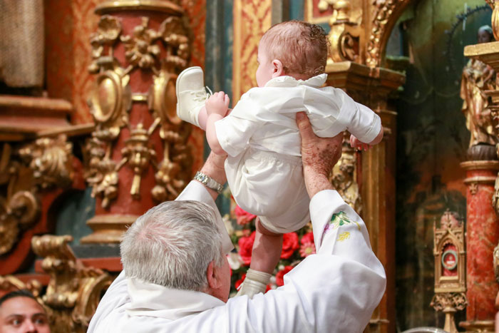 A priest baptizes a baby in white attire. This image shows a child being baptized, a key event in the MIL secret baptism story.