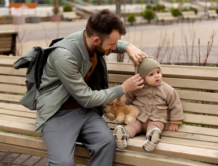 A loving dad adjusts his child's hat while sitting on a bench, a tender moment before a possible meltdown.