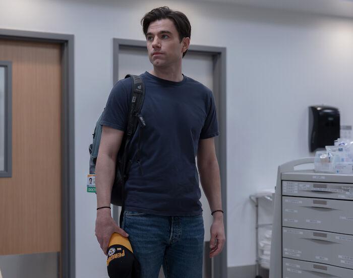 Actor Zach Cherry, wearing a navy shirt and backpack, holds a yellow cap, reflecting on a "$750 A Week" salary confession.