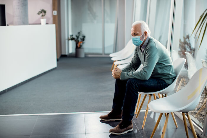An older man in a face mask sits in a doctor's waiting room, appearing to be one of the patients doctors wanted to ask about being alive right now.