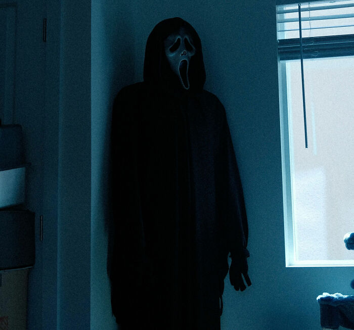 Scary figure in a dark cloak wearing a ghost face mask inside a dimly lit room