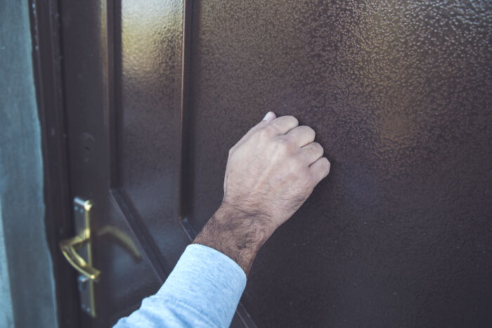 Person knocking on a brown door wearing a light blue long sleeve shirt
