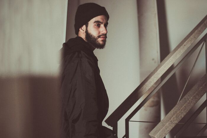 Man in dark clothes and beanie standing indoors by stairs