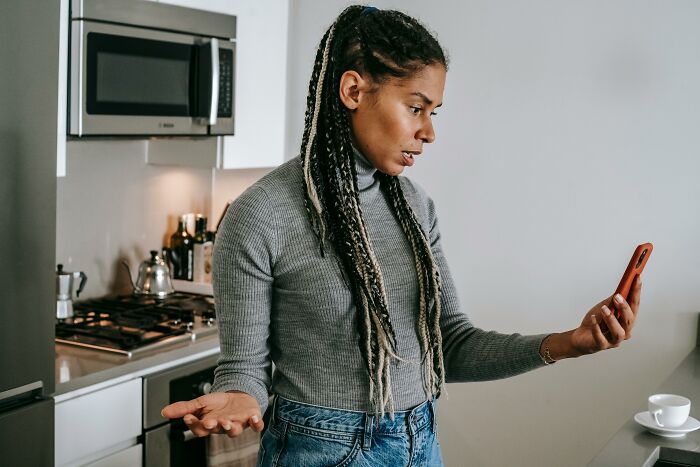 A Gen Z person with braids, looking confused at their phone in a kitchen, representing things older generations don't understand.