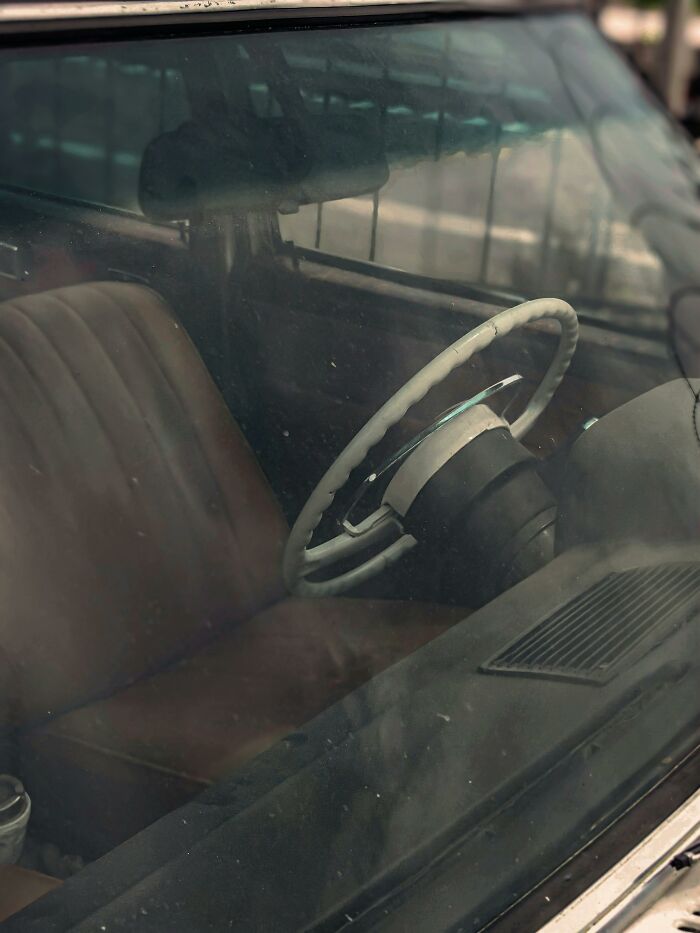 A dusty, vintage car interior through a grimy windshield. Brown leather seats and a white steering wheel. Cheating discovered.