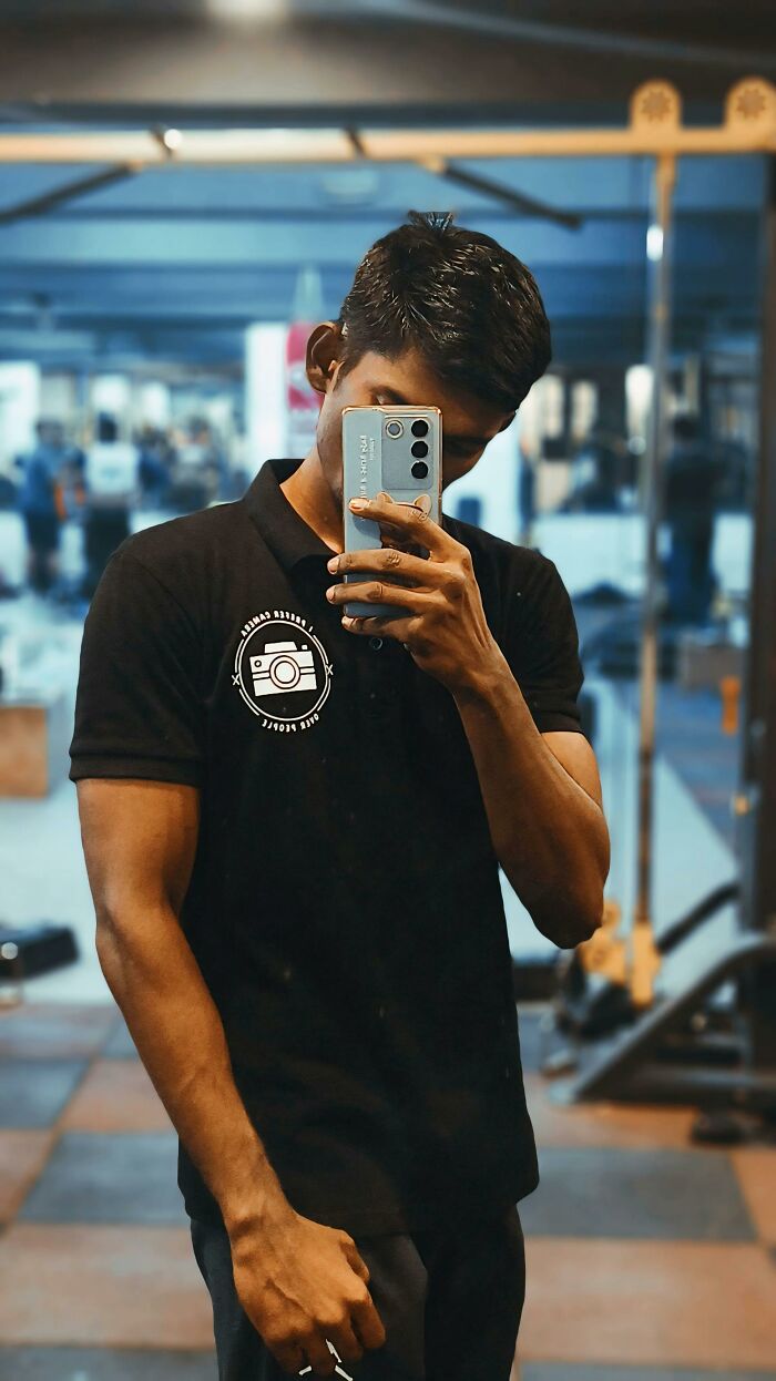 Gen Z man takes a selfie in a gym mirror, showing a phone with a unique case and a camera logo shirt.