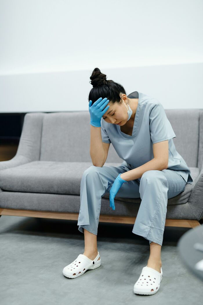 A tired nurse in blue scrubs and gloves sits on a couch, head in hand, reflecting patient notes from charts.