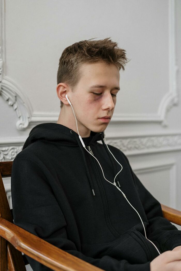 A young man with a black eye, wearing a black hoodie and earbuds, appearing troubled by deeply immoral things.