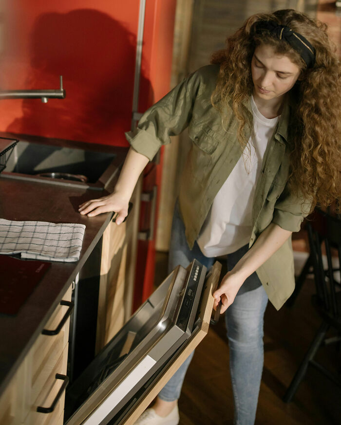 Young woman with curly hair opens a dishwasher in a kitchen, showing habits that might save you money.